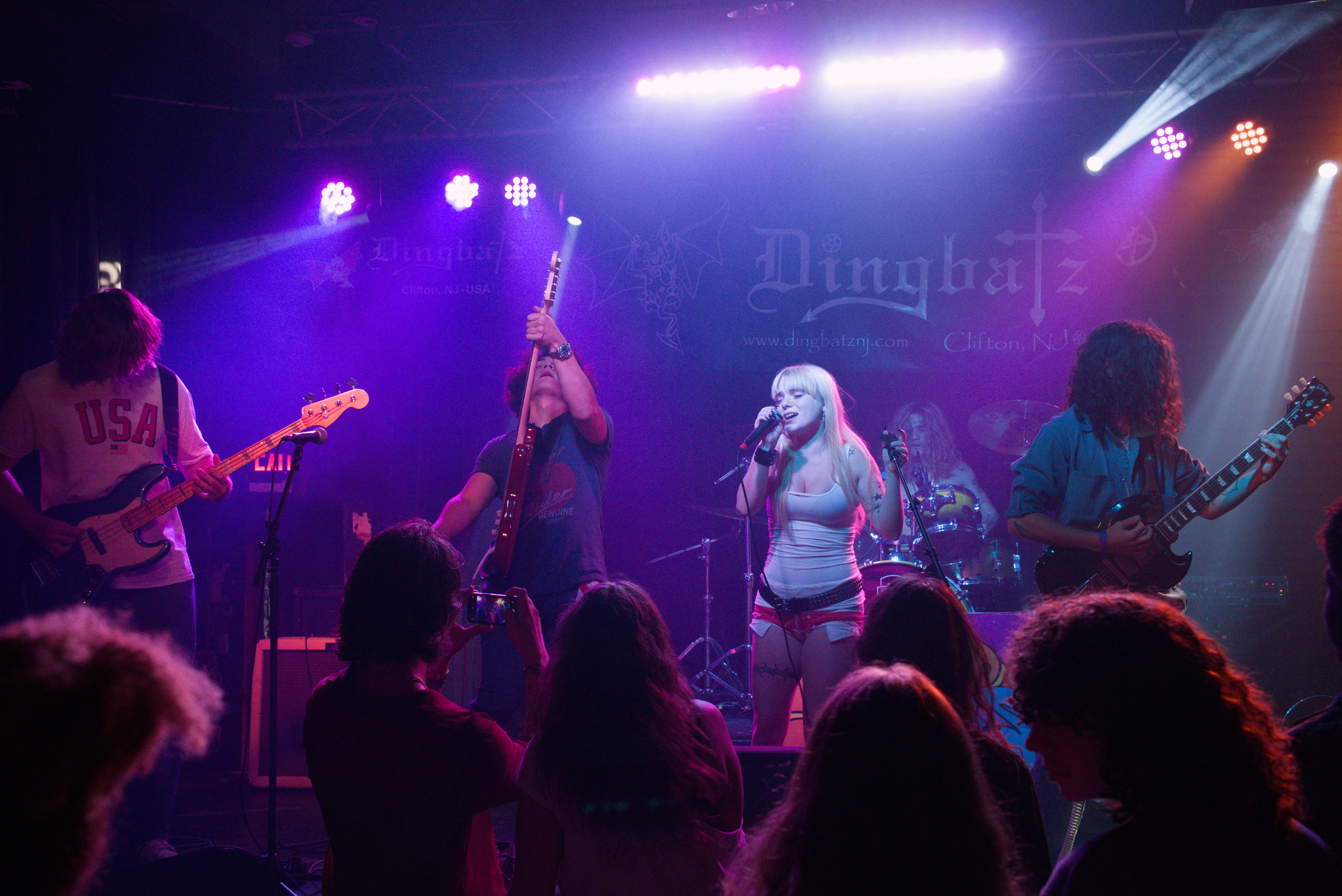 The band mid-set at Dingbatz under magenta and blue beams, the singer at the mic with her back to the crowd and the club's gothic-lettered backdrop lit behind the stage